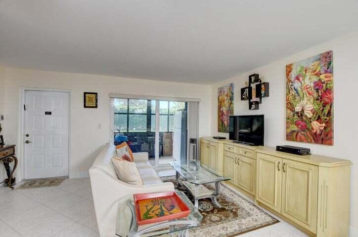 A living room features a white sofa, glass coffee table, media console, and floral wall art — 7539 S Oriole Boulevard # 103, Delray Beach FL 33446, DELRAY BEACH, FL