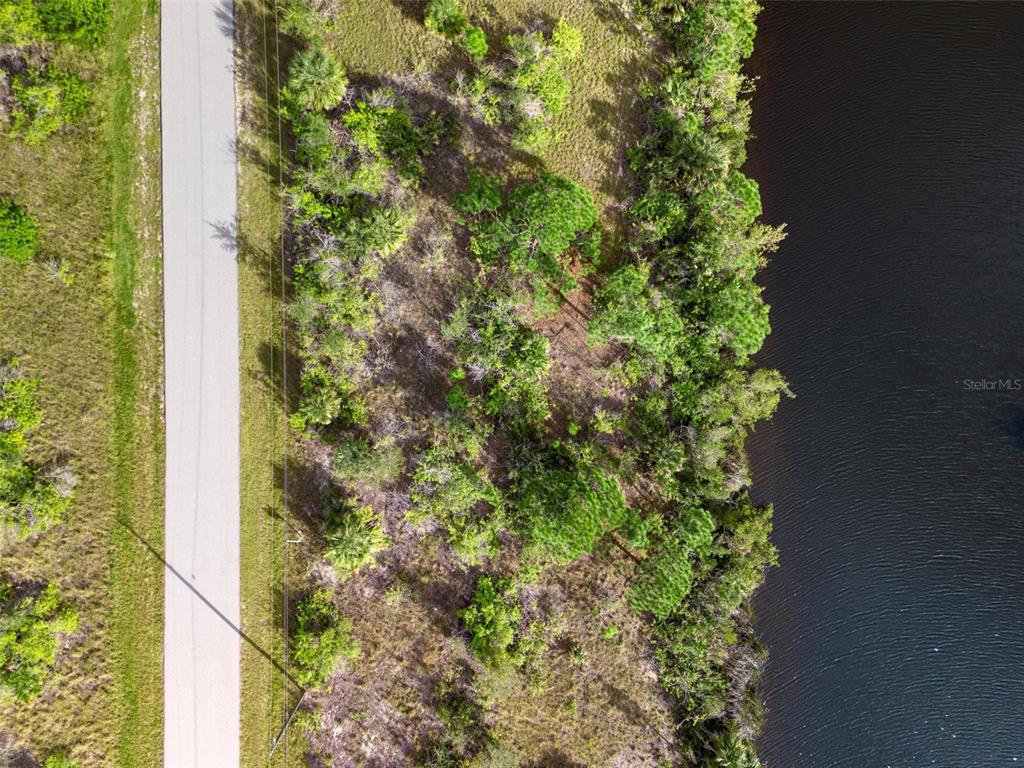 An aerial view shows a paved road running alongside a wooded lot bordering a lake — 15109 AMSTERDAM AVENUE, PORT CHARLOTTE, FL