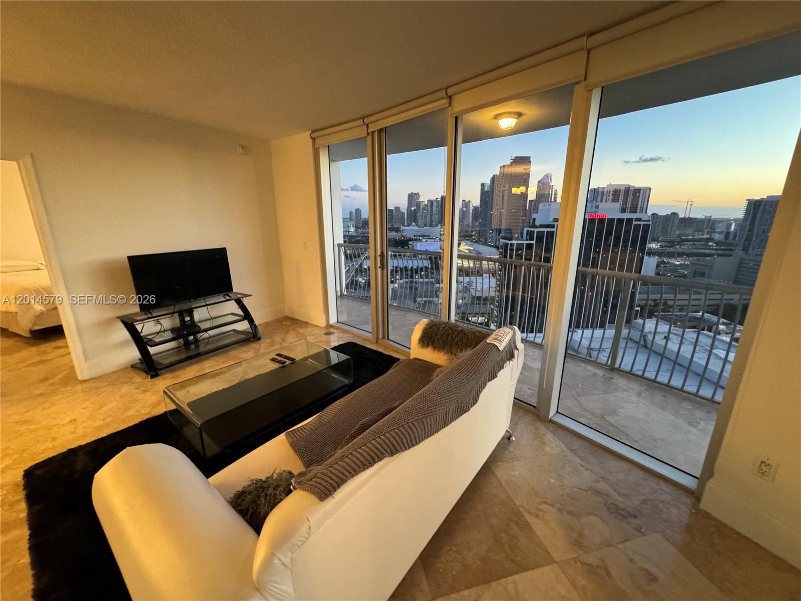 A living room with a white sofa, glass coffee table, and floor-to-ceiling windows overlooking city buildings — 1750 N Bayshore Dr # 3114, Miami FL 33132, MIAMI, FL