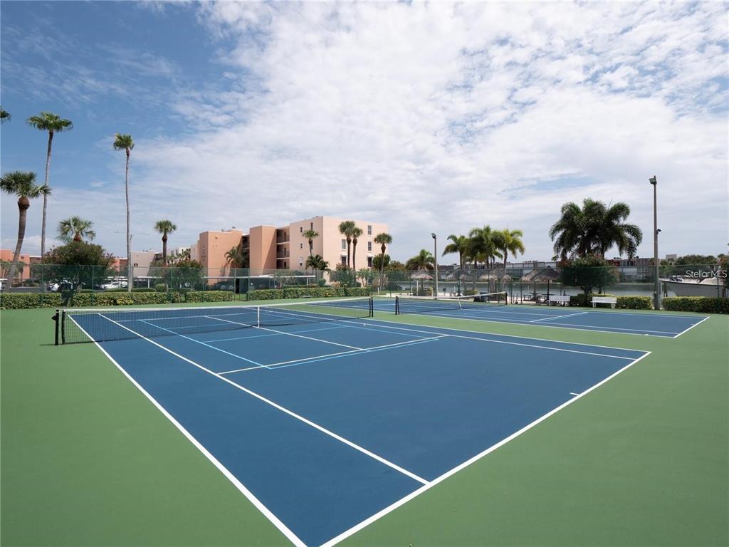 Two outdoor tennis courts with blue surfaces are surrounded by palm trees and buildings — 7405 BAY ISLAND DRIVE S UNIT 224, SOUTH PASADENA, FL