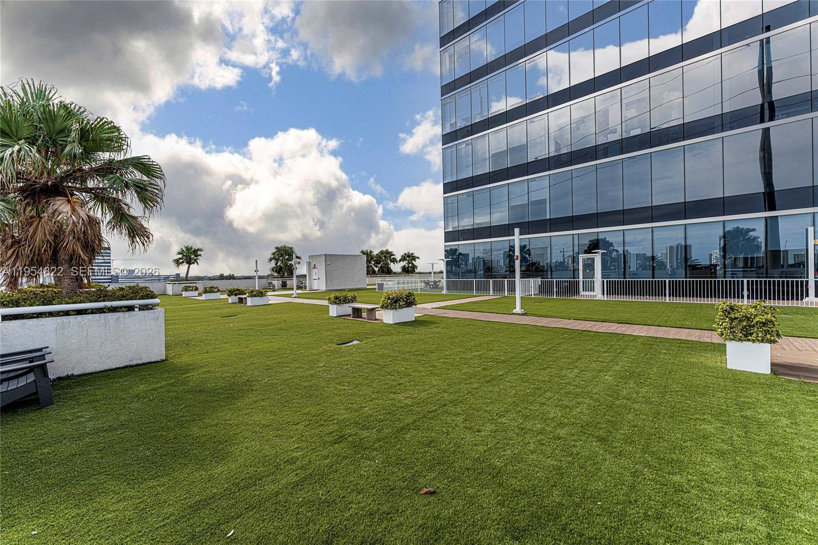 A rooftop terrace features artificial turf, potted plants, and a modern glass office building — 20200 W Dixie Hwy # 903, Miami FL 33180, MIAMI, FL
