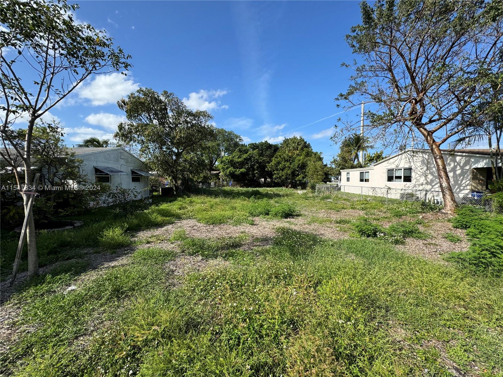 An empty, grassy lot sits between two single-story residential buildings under a clear blue sky — 2205 Coolidge St, Hollywood FL 33020, HOLLYWOOD, FL