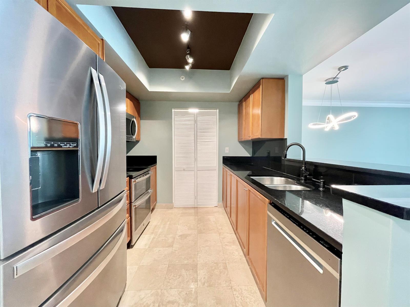 A galley kitchen features stainless steel appliances, wood cabinets, and black granite countertops — 99 SE Mizner Boulevard # 736, Boca Raton FL 33432, BOCA RATON, FL