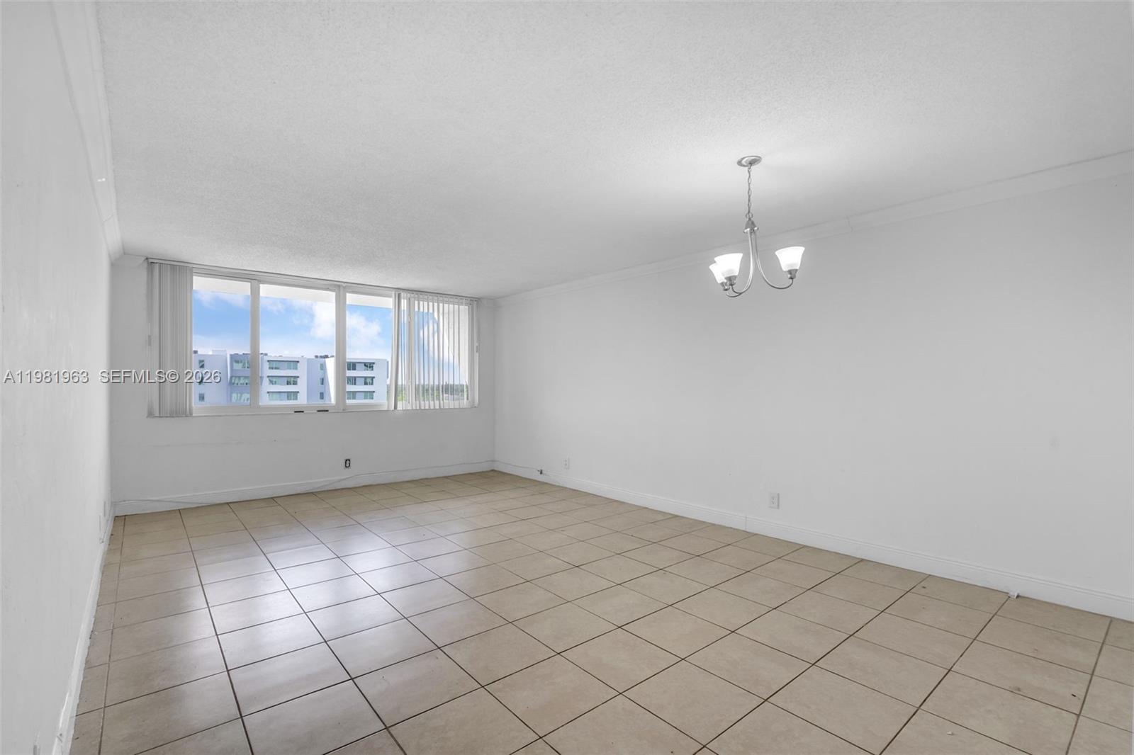 An empty room features beige tile flooring, white walls, a chandelier, and large windows — 9143 SW 77th Ave # B710, Miami FL 33156, MIAMI, FL