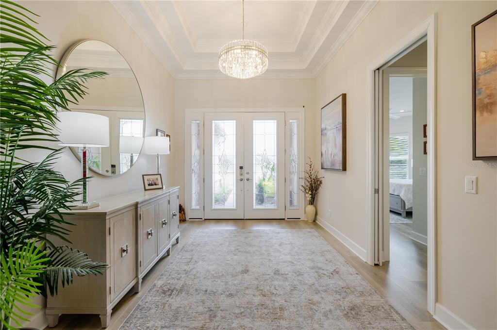A bright foyer features a tray ceiling, crystal chandelier, large round mirror, and light wood flooring — 17602 TERRACINA DRIVE, FORT MYERS, FL