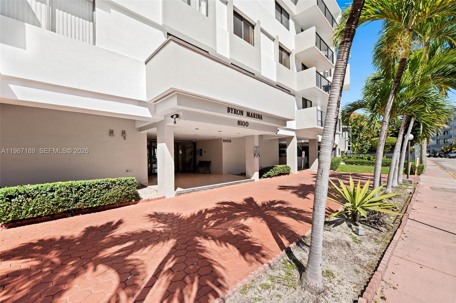 The exterior of a white multi-story building features a covered entrance with palm trees — 8100 Byron Ave # 303, Miami Beach FL 33141, MIAMI BEACH, FL