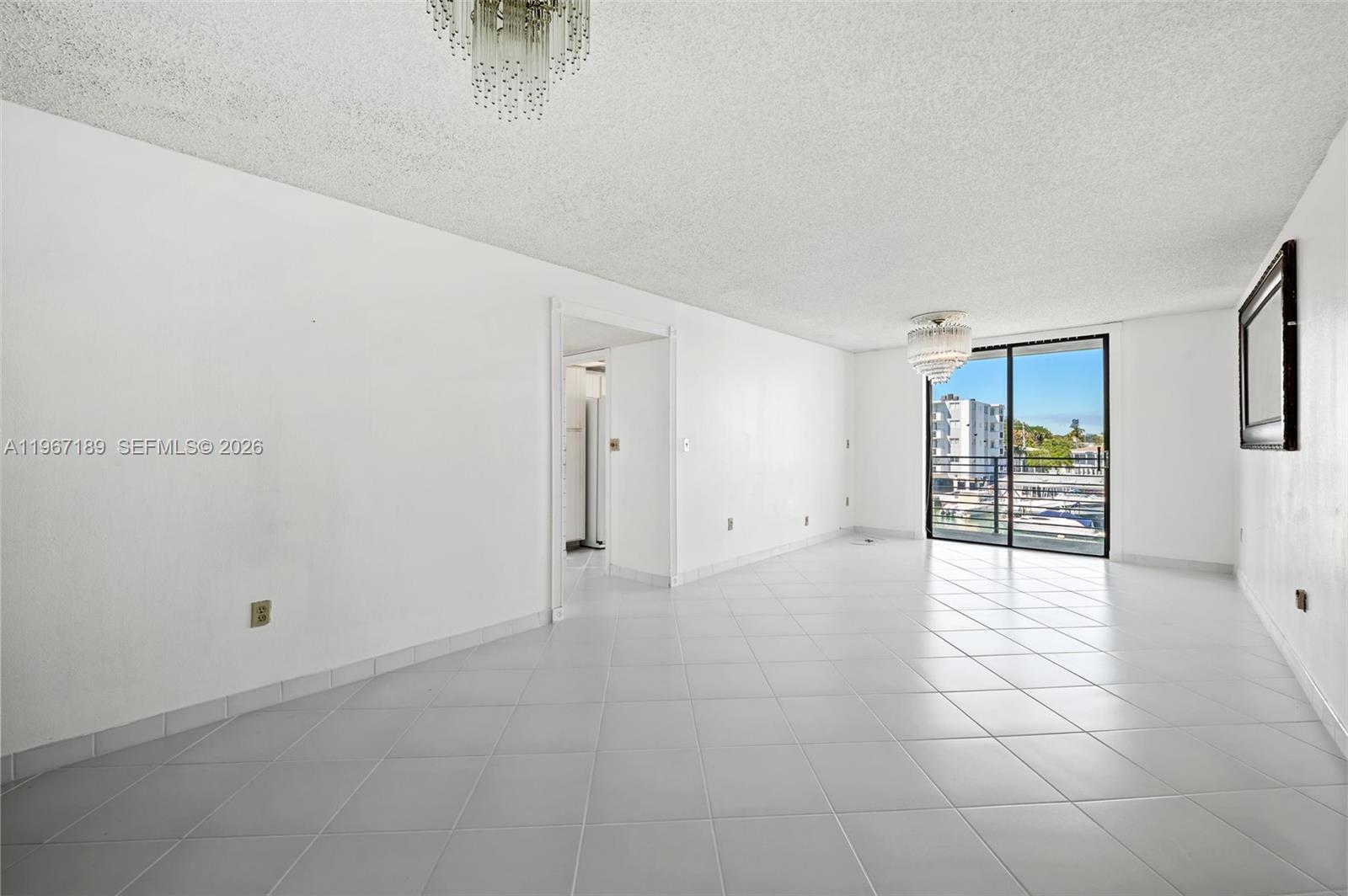 An empty living room features white tile floors, white walls, two chandeliers, and a balcony — 8100 Byron Ave # 303, Miami Beach FL 33141, MIAMI BEACH, FL