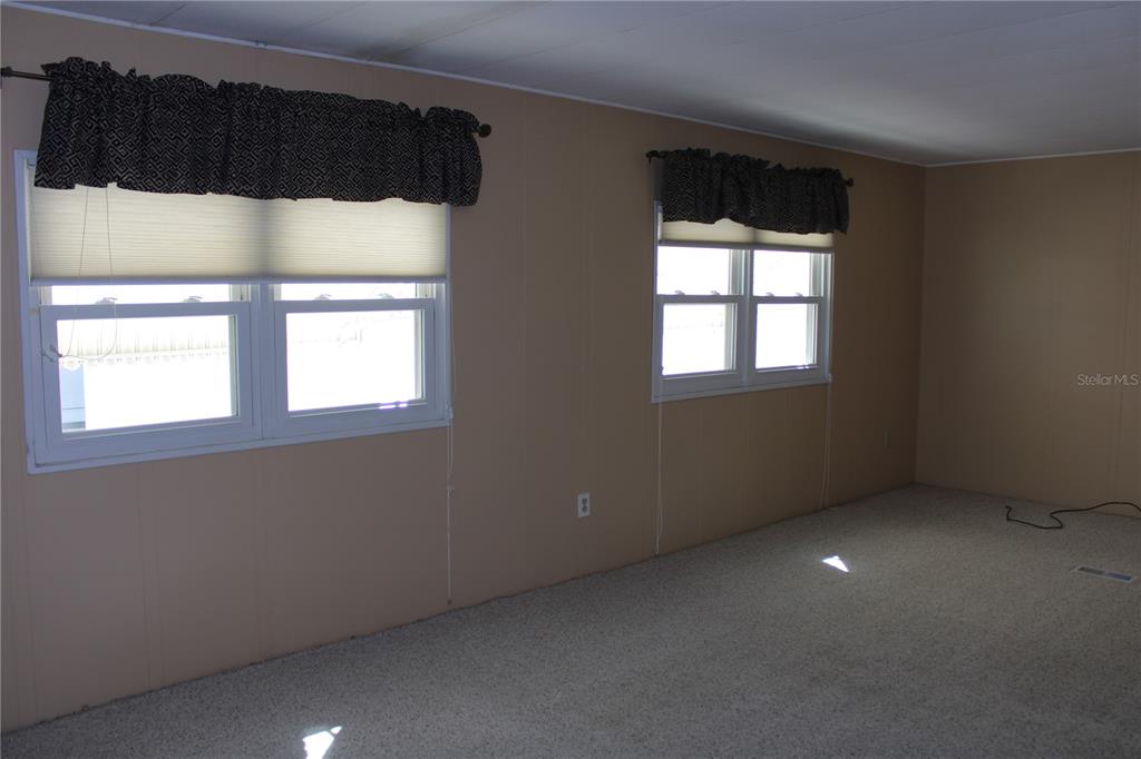 An empty room features beige paneled walls, beige carpeting, and two windows with valances — 2107 PALMA SOLA BOULEVARD UNIT 8, BRADENTON, FL