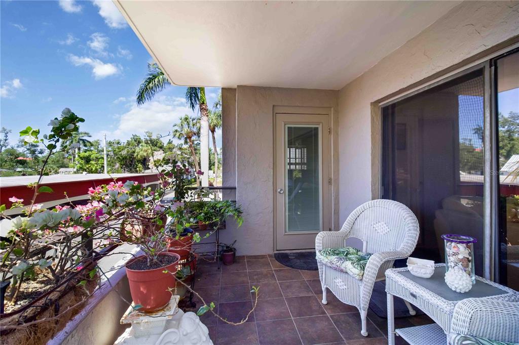 A tiled balcony features a white wicker chair, a small table, and several potted plants — 1400 BARCARROTA BOULEVARD UNIT 201, BRADENTON, FL