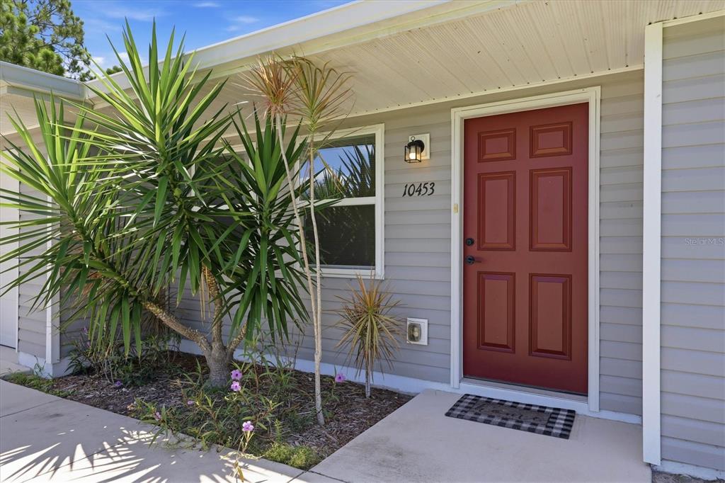 A red front door with a house number sits beside a window and tropical plants — 10453 REIMS AVENUE, ENGLEWOOD, FL