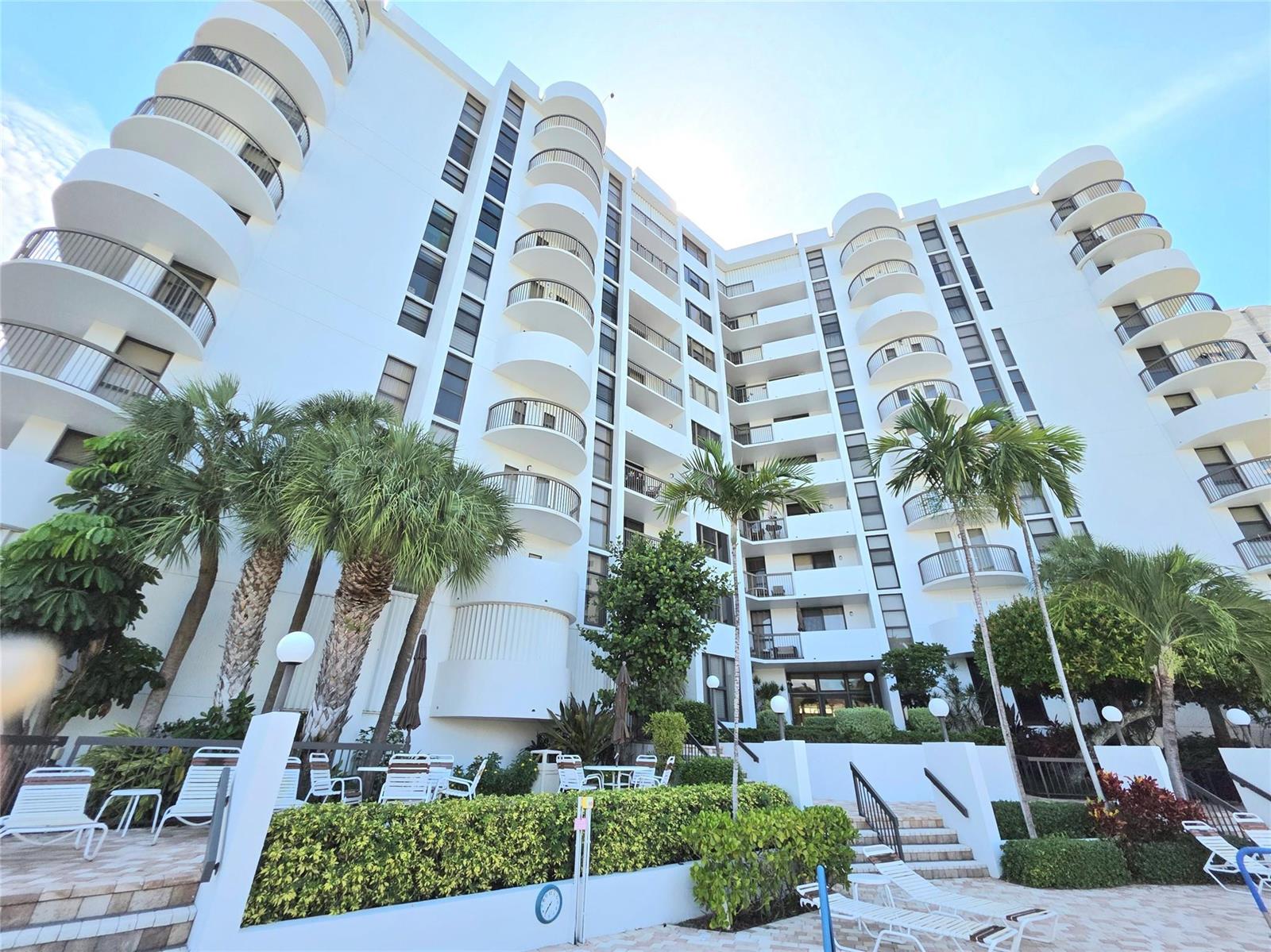 A low-angle view of a white multi-story apartment building with curved balconies and palm trees — 1631 S Ocean Boulevard # 1006, Pompano Beach FL 33062, POMPANO BEACH, FL