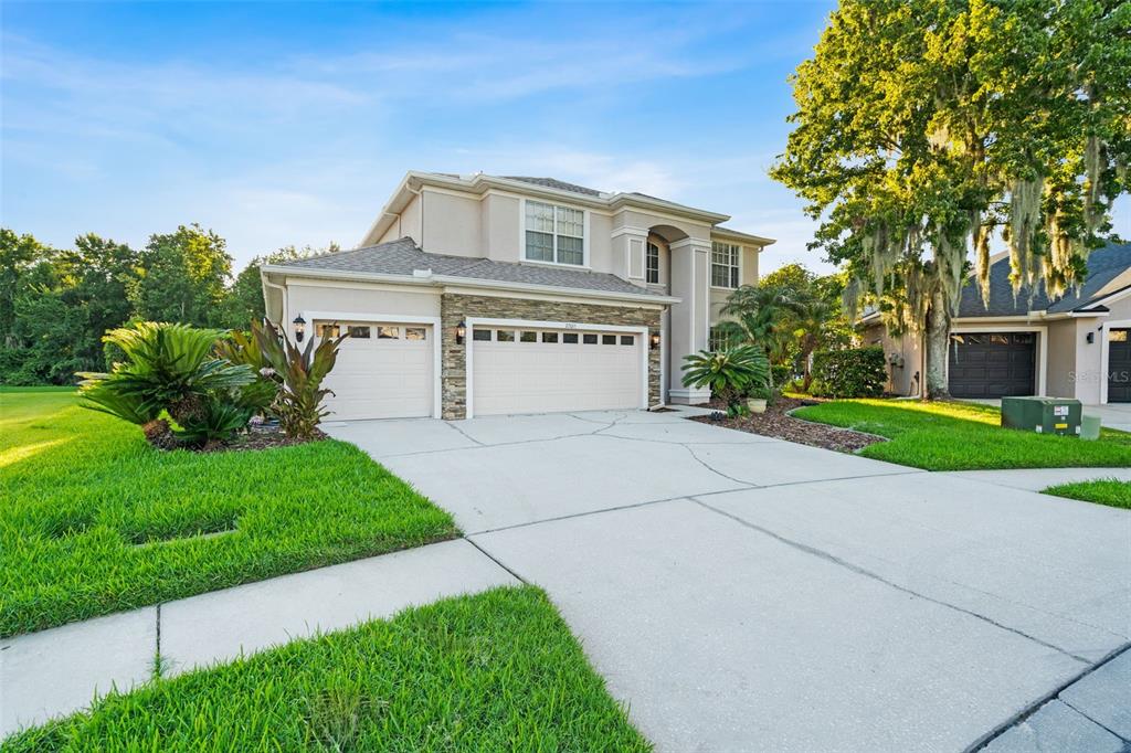 A two-story house with a three-car garage and stone facade sits on a grassy lot — 27015 FIREBUSH DRIVE, WESLEY CHAPEL, FL