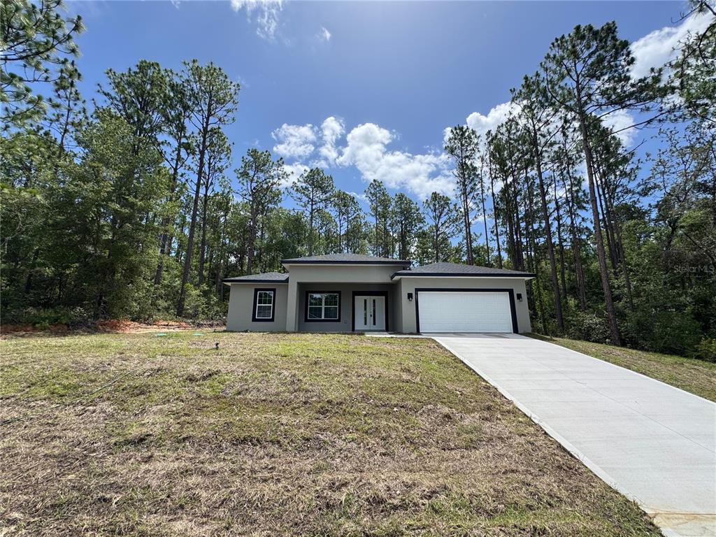 A single-story house with a white garage and concrete driveway sits in a wooded lot — 7687 N QUIBBLE AVENUE, DUNNELLON, FL