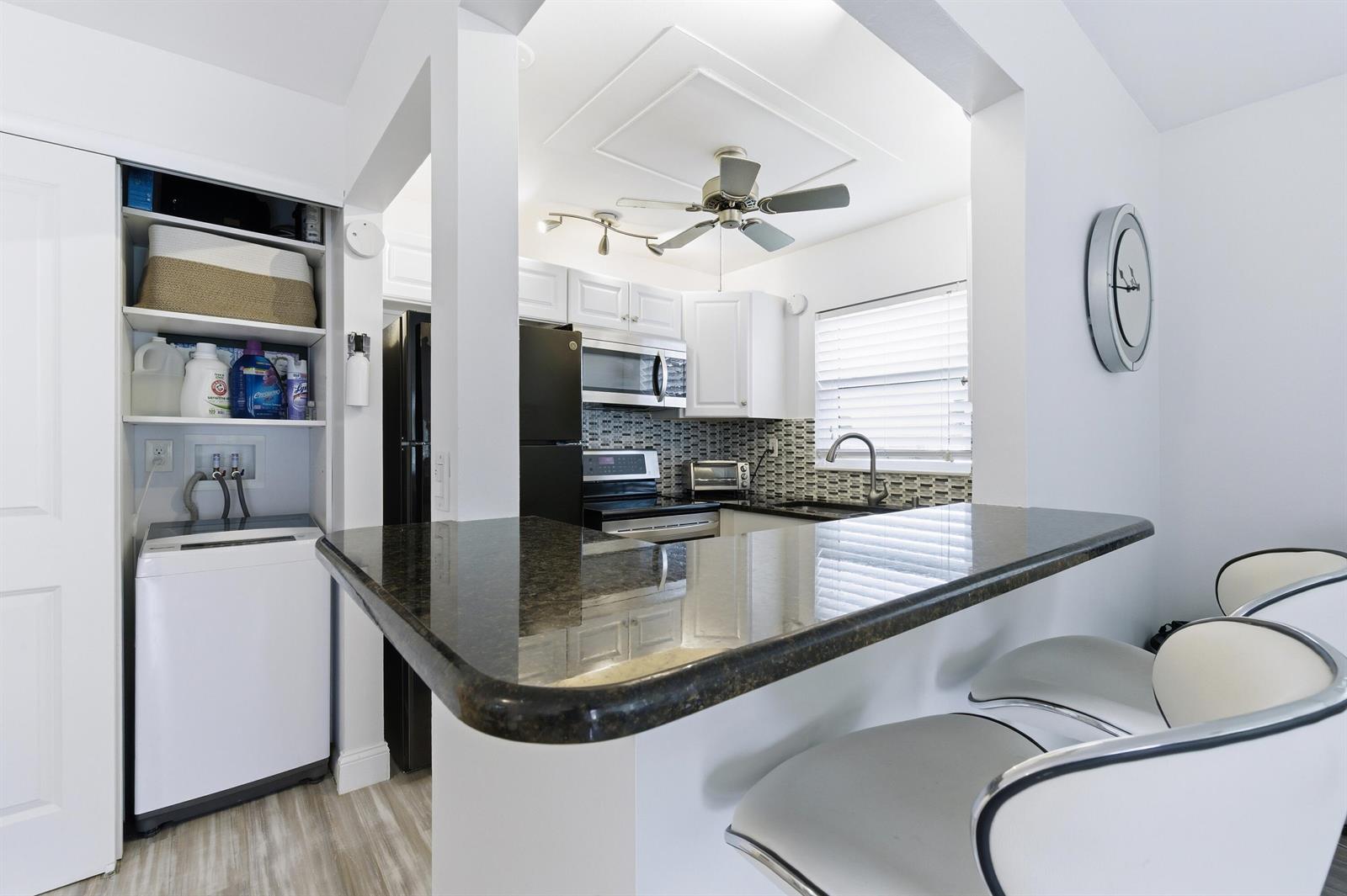 A kitchen features a granite breakfast bar, white cabinetry, black appliances, and an adjacent laundry area — 70 Bedford # C, West Palm Beach FL 33417, WEST PALM BEACH, FL