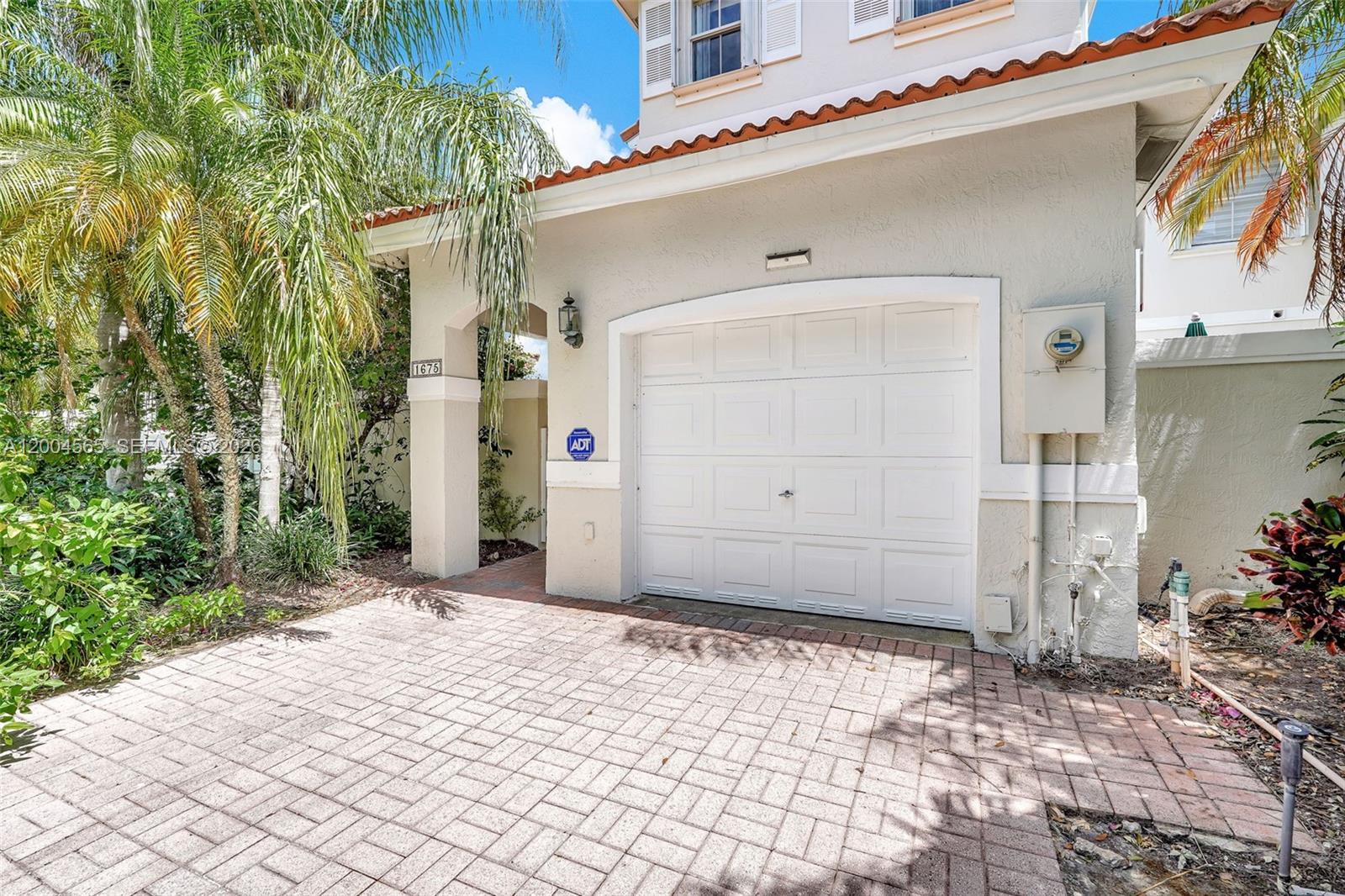 A white garage door is set into a beige stucco home with a brick driveway — 1675 Yellowheart Way, Hollywood FL 33019, HOLLYWOOD, FL