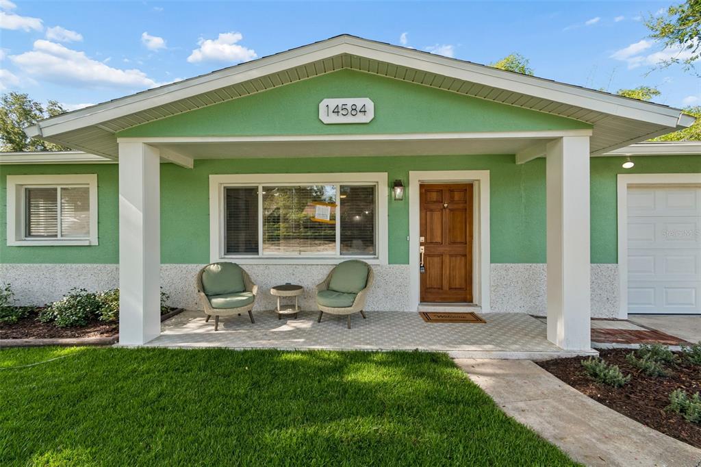 A green house exterior features a covered porch with two chairs and a wooden door — 14584 APACHE AVENUE, LARGO, FL