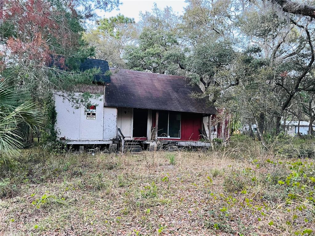 A weathered, single-story house with a dark roof stands amidst overgrown vegetation and trees — 19435 SE 92ND PLACE, OCKLAWAHA, FL