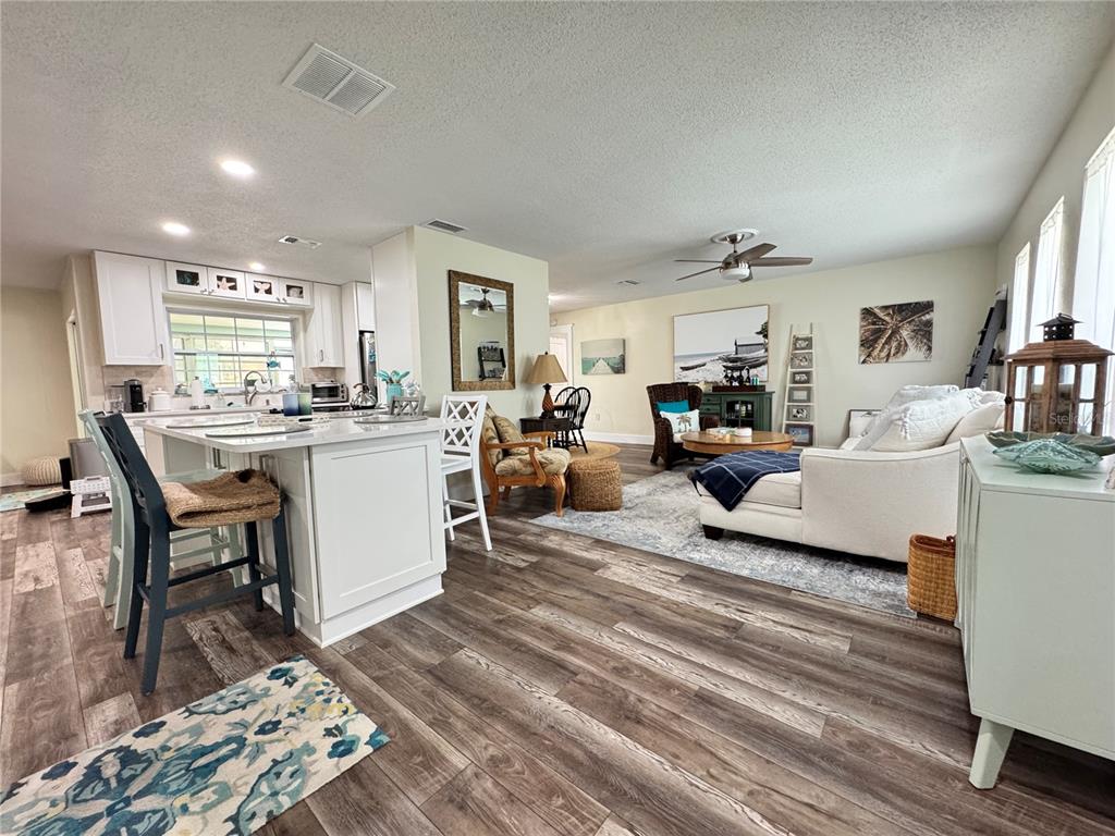 An open-concept kitchen and living area features wood-look flooring, white cabinetry, and neutral furniture — 6813 13TH AVENUE DRIVE W, BRADENTON, FL