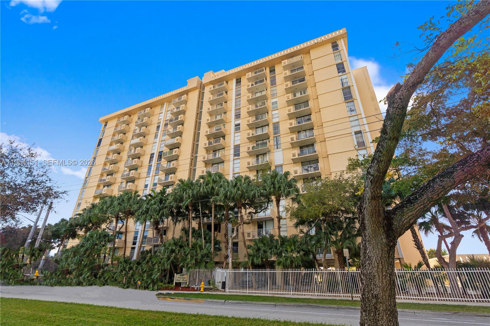 A multi-story tan apartment building stands behind a white fence and palm trees — 2350 NE 135th St # 1202, North Miami FL 33181, NORTH MIAMI, FL