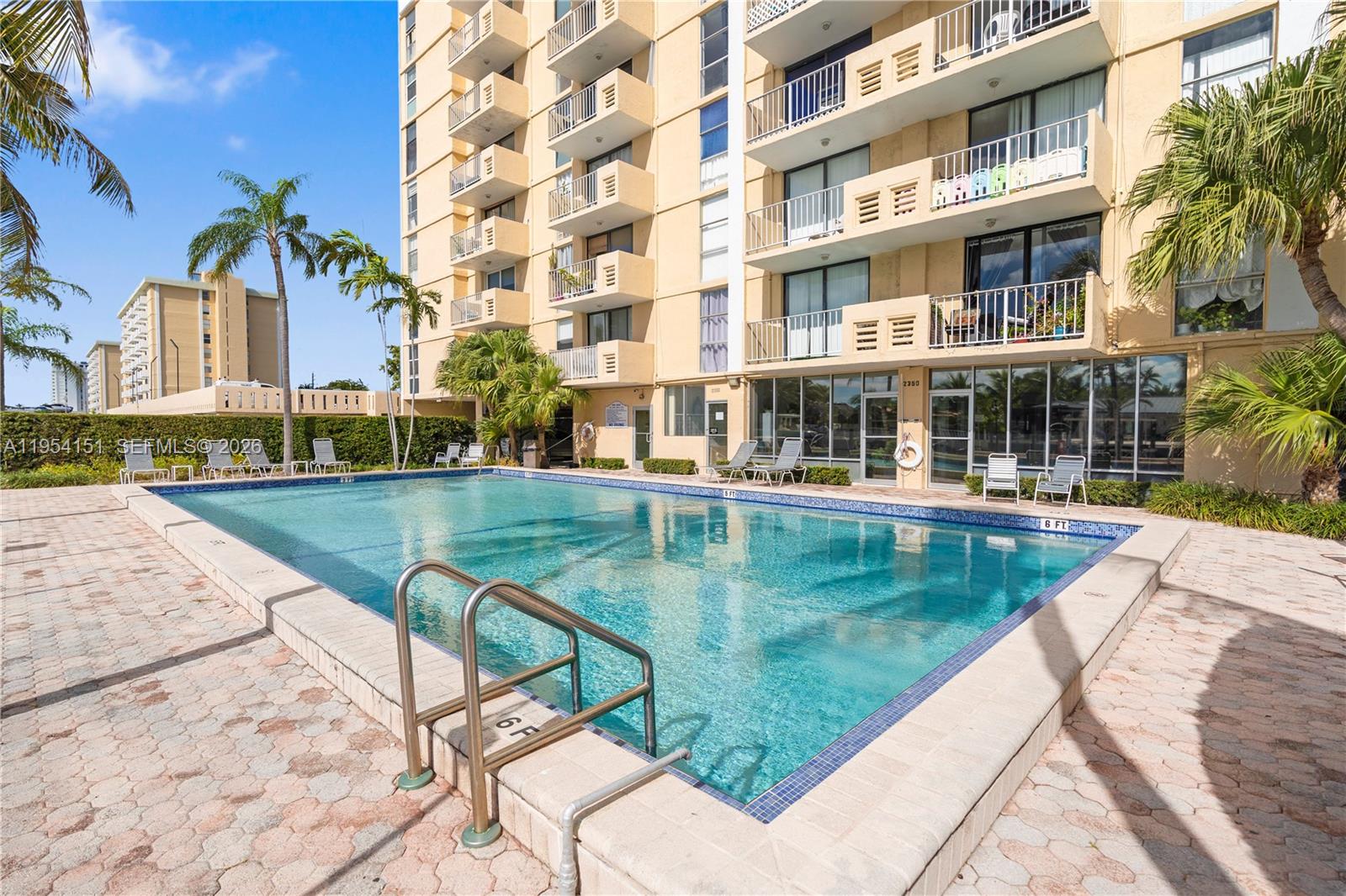 A rectangular swimming pool sits on a paved deck in front of a multi-story apartment building — 2350 NE 135th St # 1202, North Miami FL 33181, NORTH MIAMI, FL