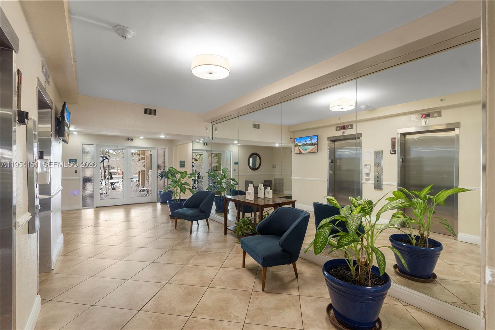 A lobby area features tile floors, blue armchairs, potted plants, and mirrored walls with elevators — 2350 NE 135th St # 1202, North Miami FL 33181, NORTH MIAMI, FL