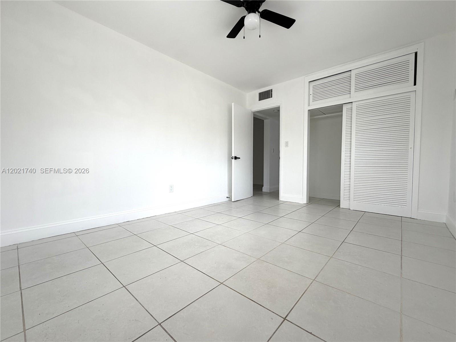 A bright, empty bedroom features white walls, light-colored tile flooring, and a louvered closet — NORTH MIAMI BEACH, FL