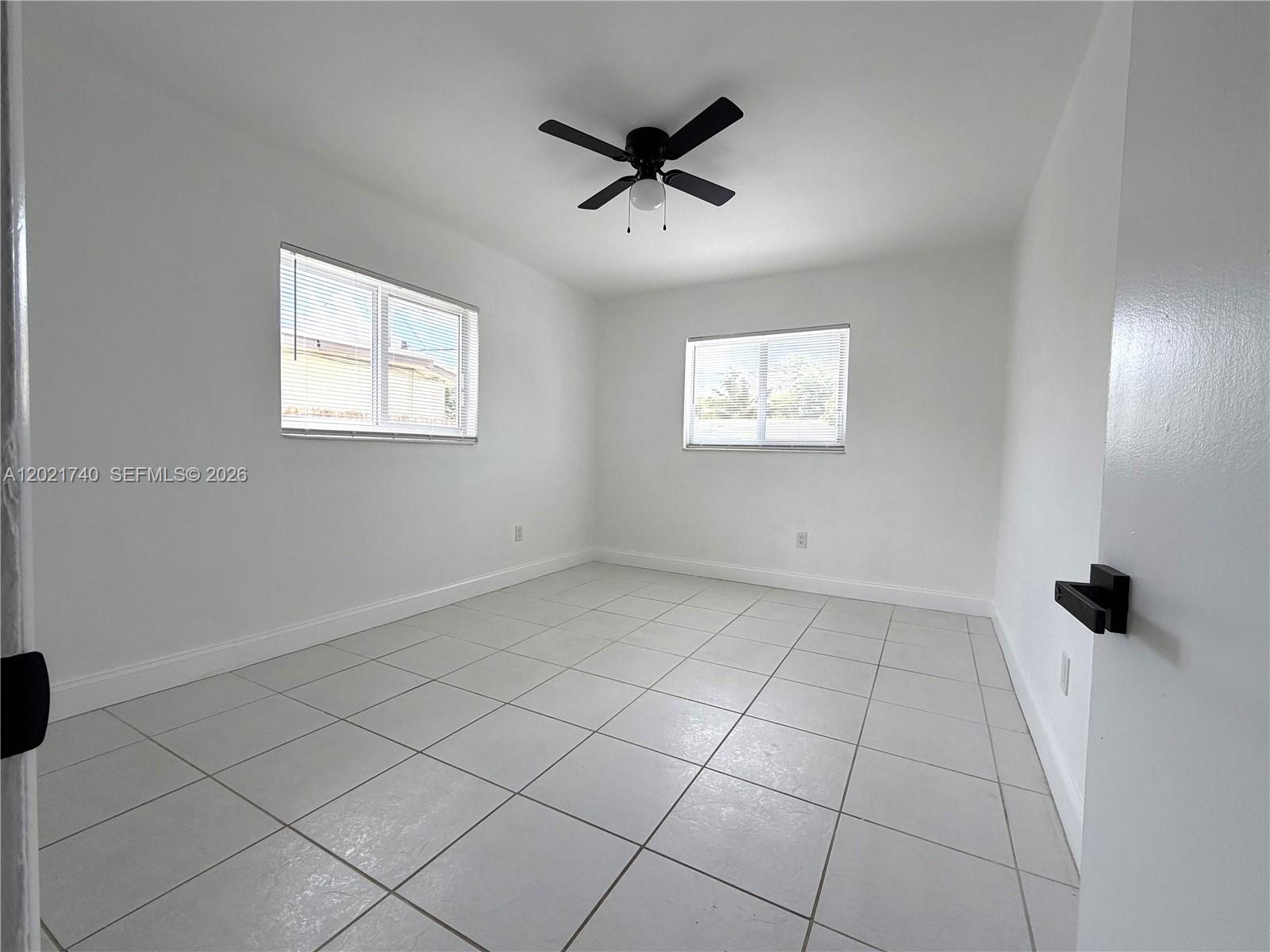 An empty bedroom features white walls, light-colored tile flooring, two windows, and a ceiling fan — NORTH MIAMI BEACH, FL
