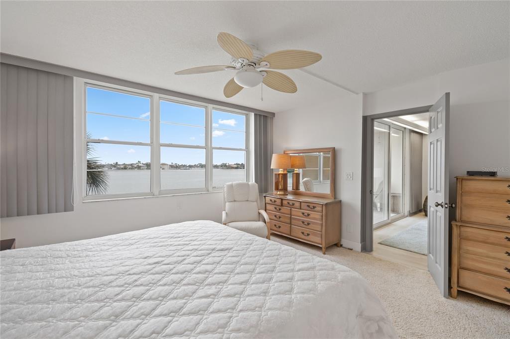 A bedroom features a white bed, a wooden dresser, and large windows overlooking the water — 7910 SUN ISLAND DRIVE S UNIT 302, SOUTH PASADENA, FL