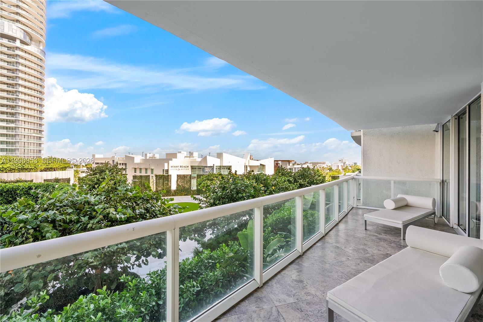 A balcony with stone flooring, glass railings, and two white lounge chairs overlooks trees — 450 Alton Rd # 704, Miami Beach FL 33139, MIAMI BEACH, FL