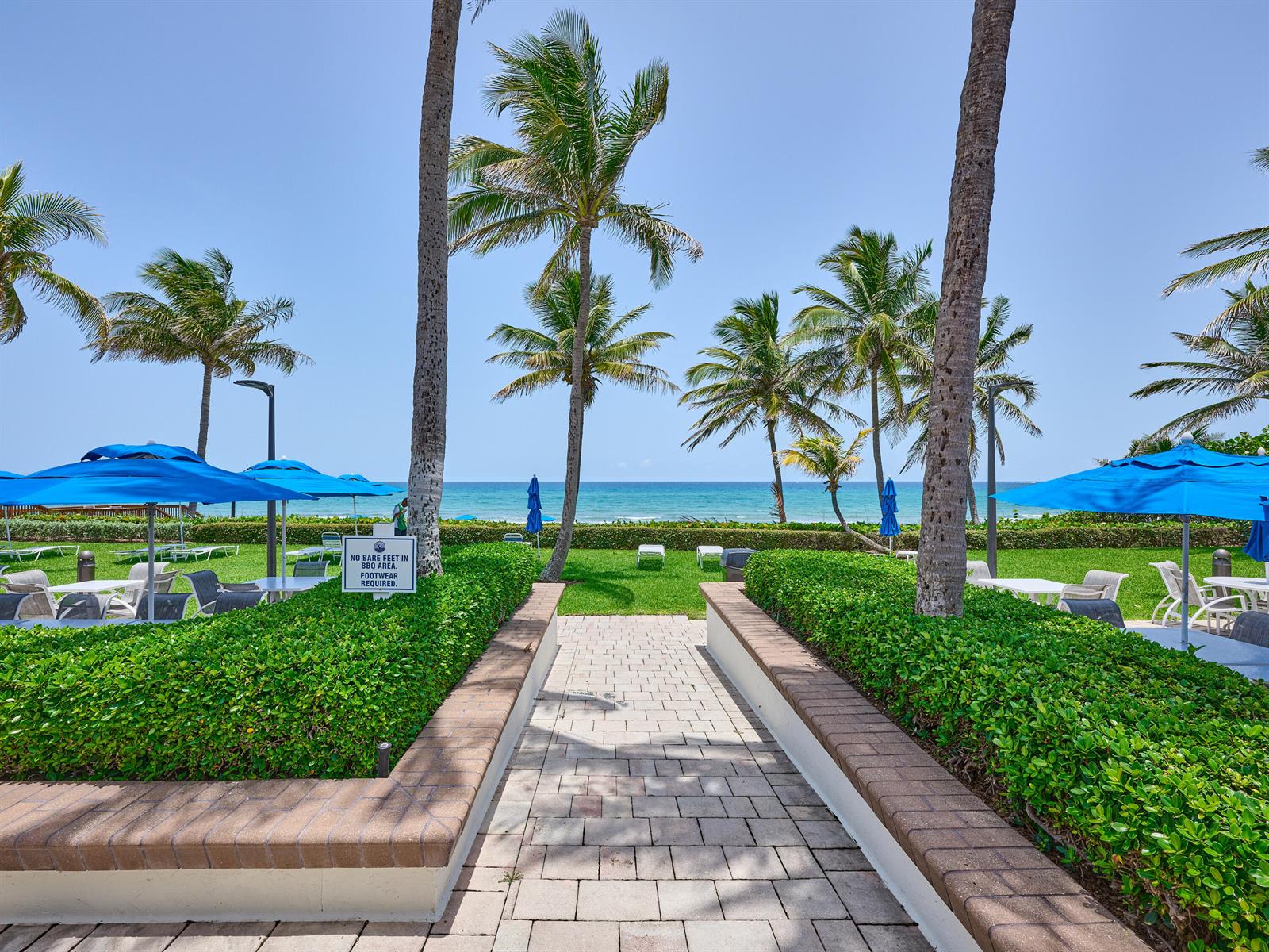 A paved walkway leads through a landscaped courtyard toward the ocean with blue patio umbrellas — 3420 S Ocean Boulevard # 8w, Highland Beach FL 33487, HIGHLAND BEACH, FL