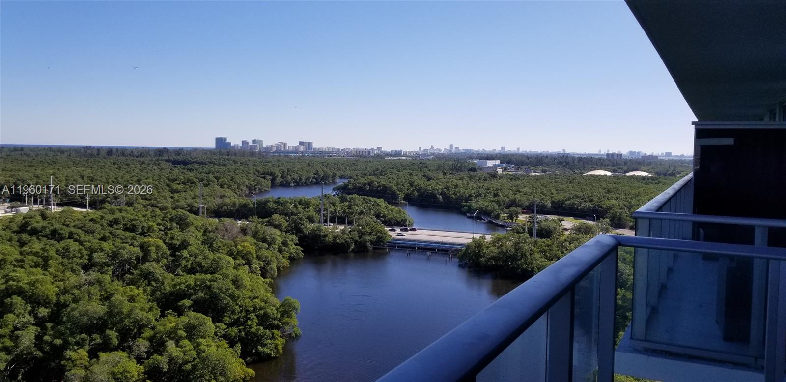 A high-angle view from a balcony overlooking a dense forest, a waterway, and a distant city — 16385 Biscayne Blvd # 1516, Miami FL 33160, MIAMI, FL