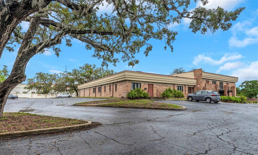 A low-rise brick commercial building sits behind a large parking lot under a sprawling tree — 10320 N 56TH STREET UNIT 100, TEMPLE TERRACE, FL