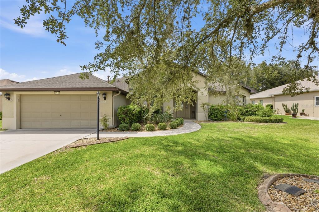 A single-story house with a beige stucco exterior, a two-car garage, and a large oak tree — 27127 STONEY BROOK DRIVE, LEESBURG, FL