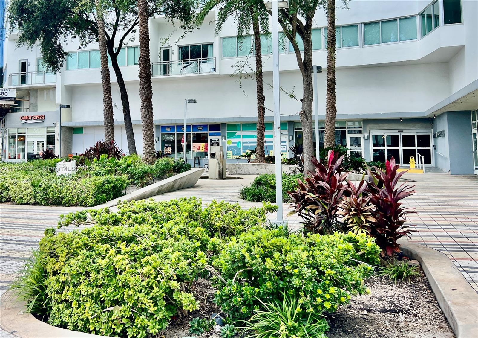 A landscaped courtyard with palm trees and shrubs sits in front of a modern building — 7135 Collins Ave # 621, Miami Beach FL 33141, MIAMI BEACH, FL
