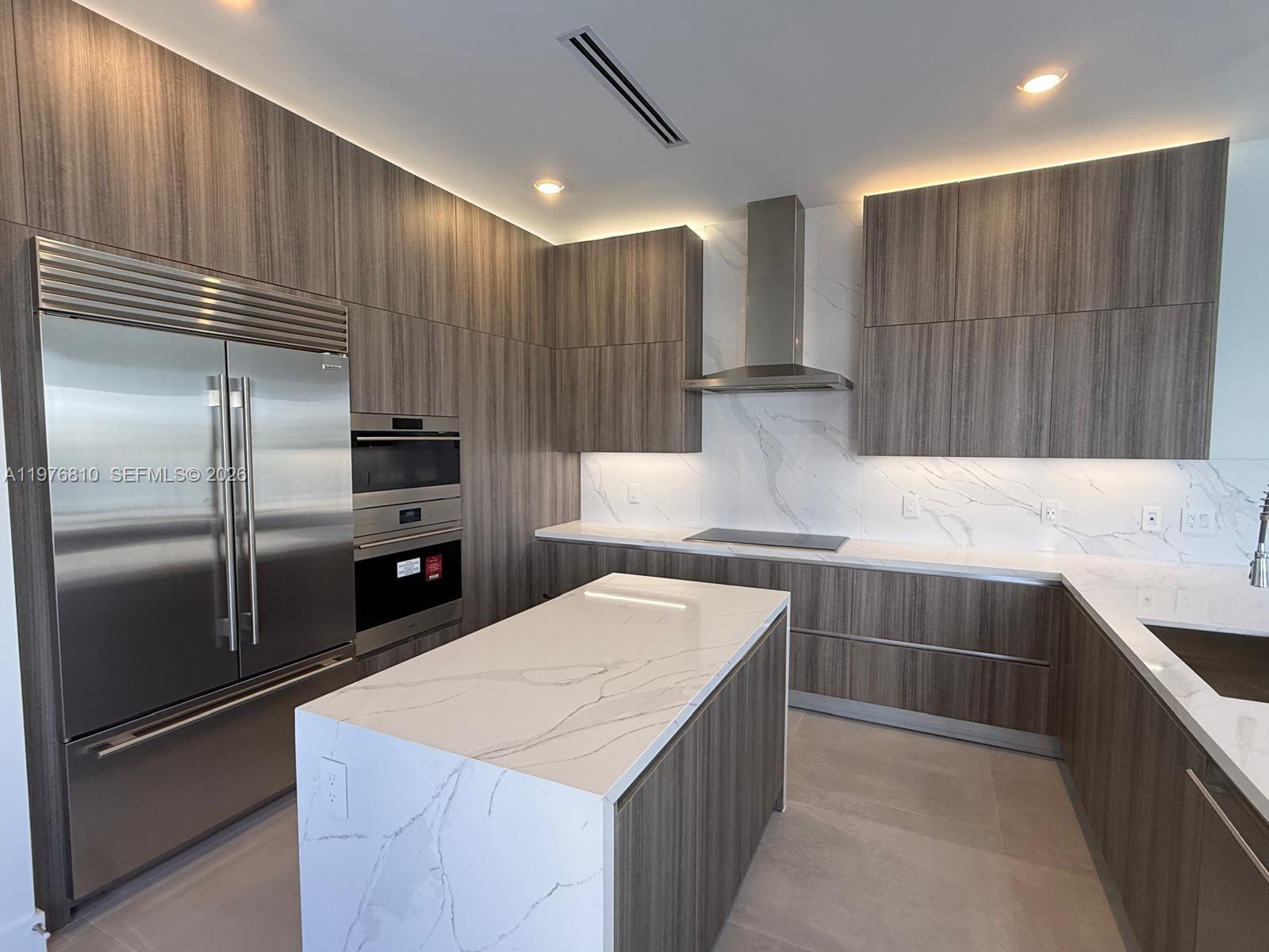 A modern kitchen features wood-grain cabinetry, a marble-topped island, and stainless steel appliances — 19616 NE 14 CT, Miami FL 33179, MIAMI, FL