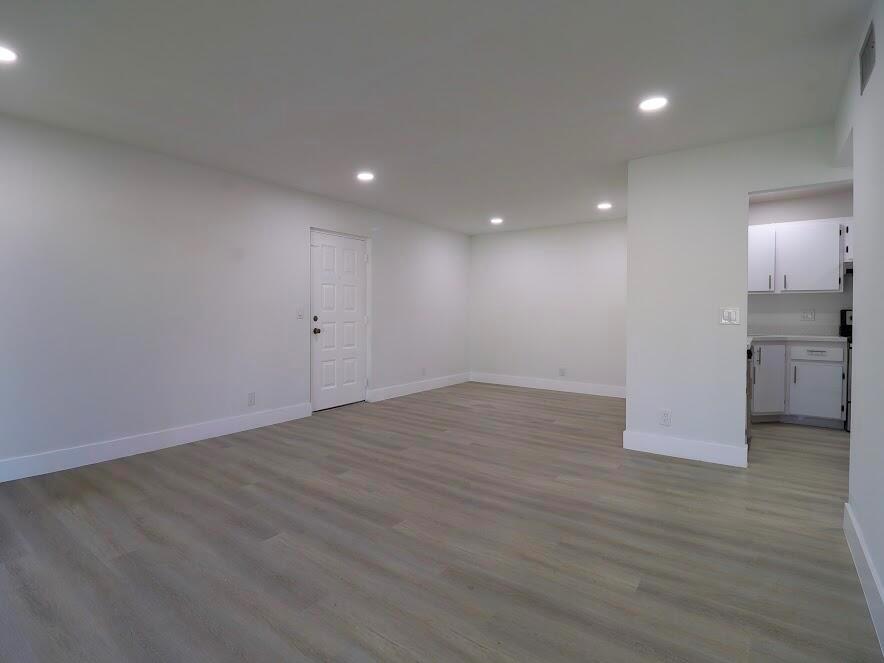 A bright, empty living room with light wood-look flooring and recessed lighting leads to a kitchen — 614 Sea Pine Way # H2, FL 33415, FL