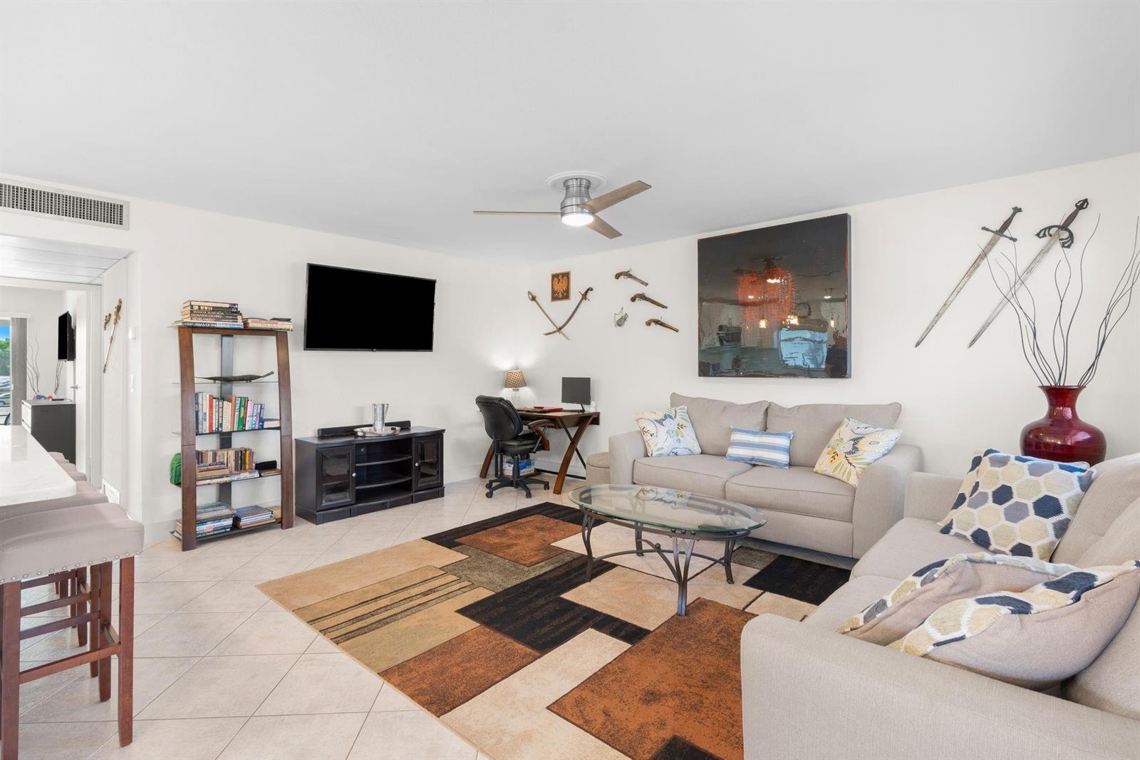 A living room features a beige sofa, a patterned area rug, and wall-mounted decorative swords — 189 Brittany D # 189, Delray Beach FL 33446, DELRAY BEACH, FL