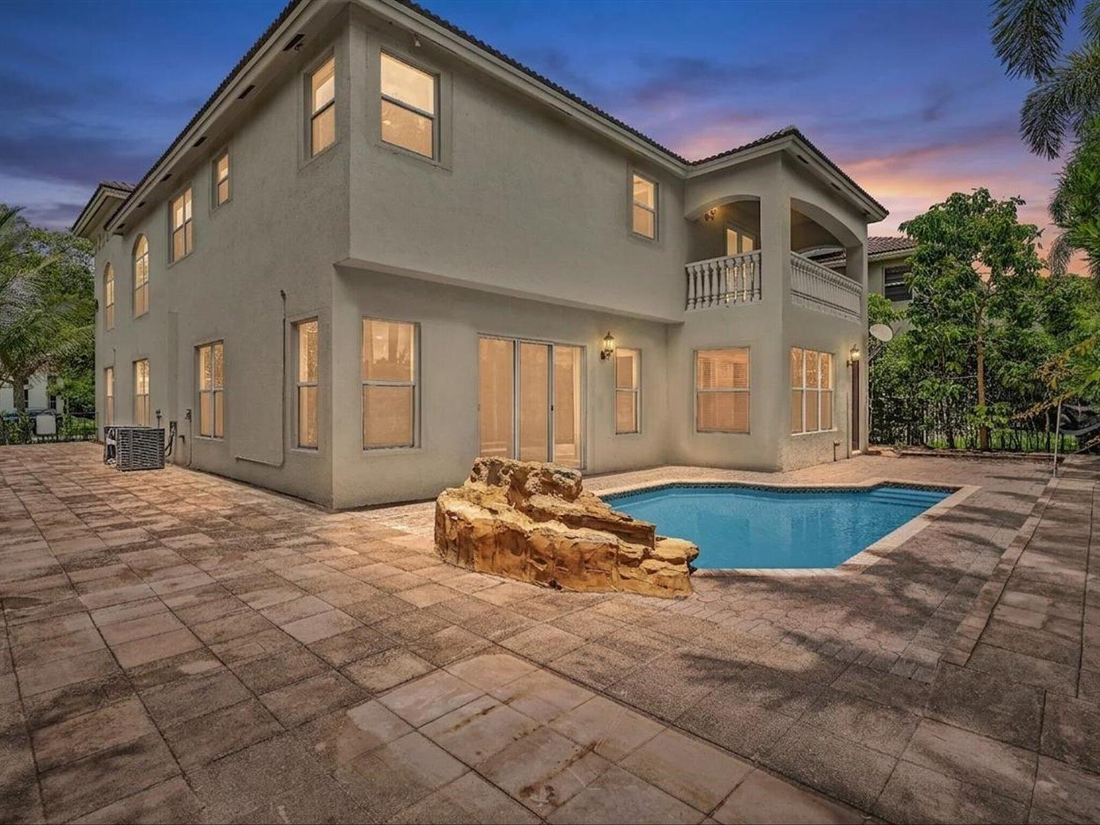 A two-story house with a backyard pool, stone feature, and paved patio at dusk — 9286 Nugent Trail, West Palm Beach FL 33411, WEST PALM BEACH, FL