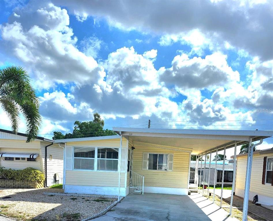 A yellow mobile home features a white carport and a gravel front yard under clouds — 6509 OREGON STREET, BRADENTON, FL