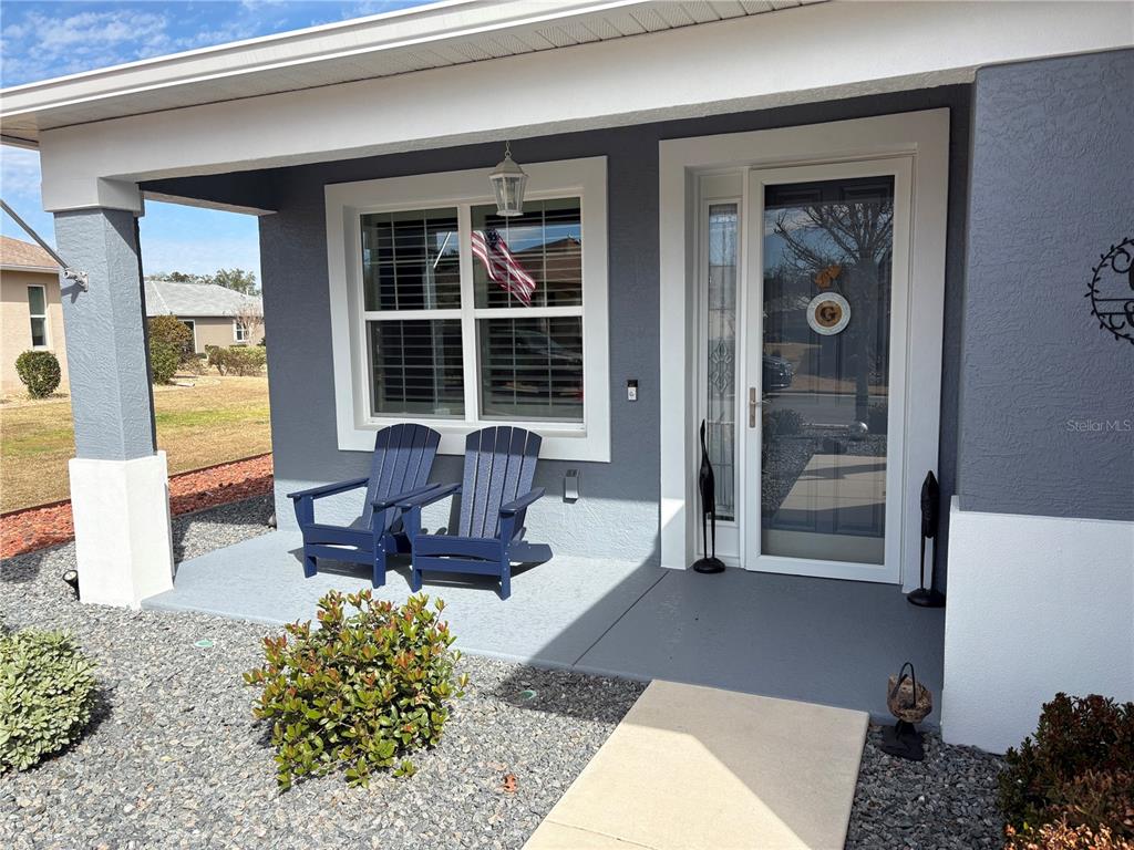 A front porch with two blue Adirondack chairs, a glass storm door, and gray siding — 7819 SW 88TH STREET ROAD, OCALA, FL