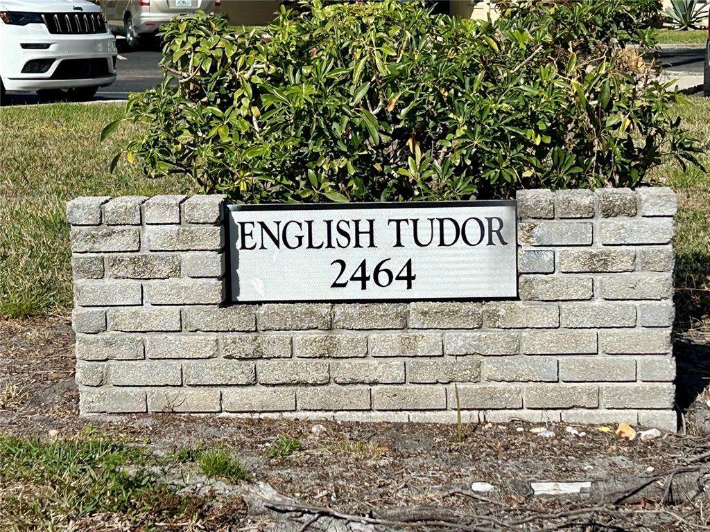 A brick wall displays a sign reading English Tudor 2464 in front of a green bush — 2464 AUSTRALIA WAY E UNIT 78, CLEARWATER, FL