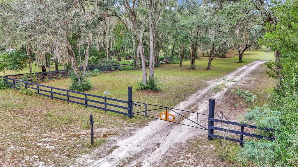 A dirt driveway leads through a black metal gate into a wooded, fenced grassy lot — 19597 SE 70TH LANE, MORRISTON, FL