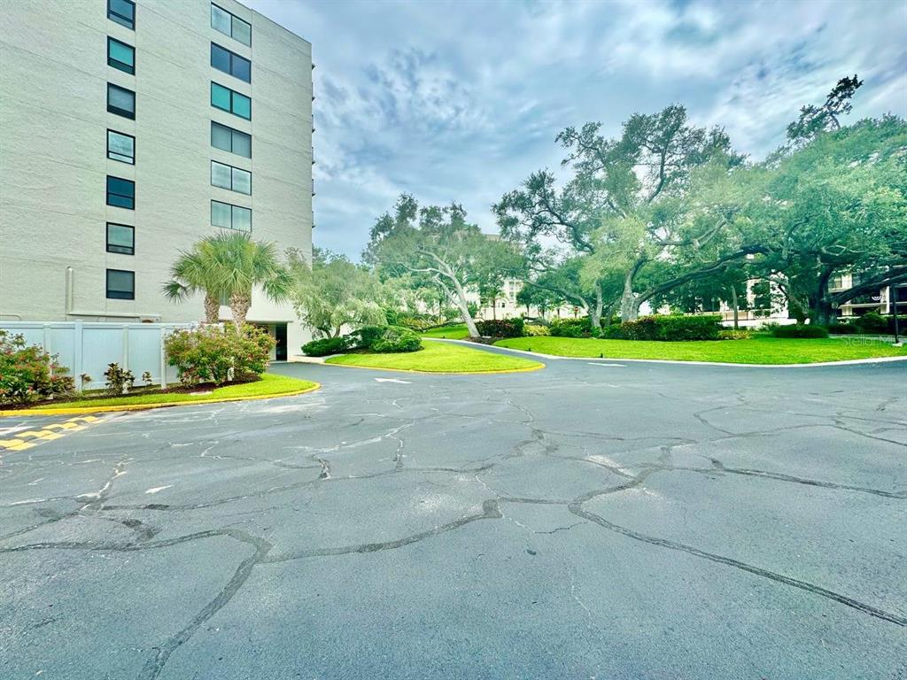 A paved parking lot sits in front of a tall building and large oak trees — 220 BELLEVIEW BOULEVARD UNIT 311, CLEARWATER, FL