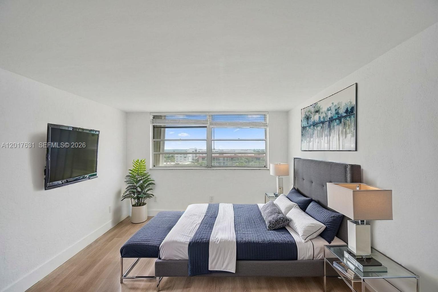 A bedroom features a gray upholstered bed, a wall-mounted television, and a large window — 7850 Byron Ave # 702, Miami Beach FL 33141, MIAMI BEACH, FL
