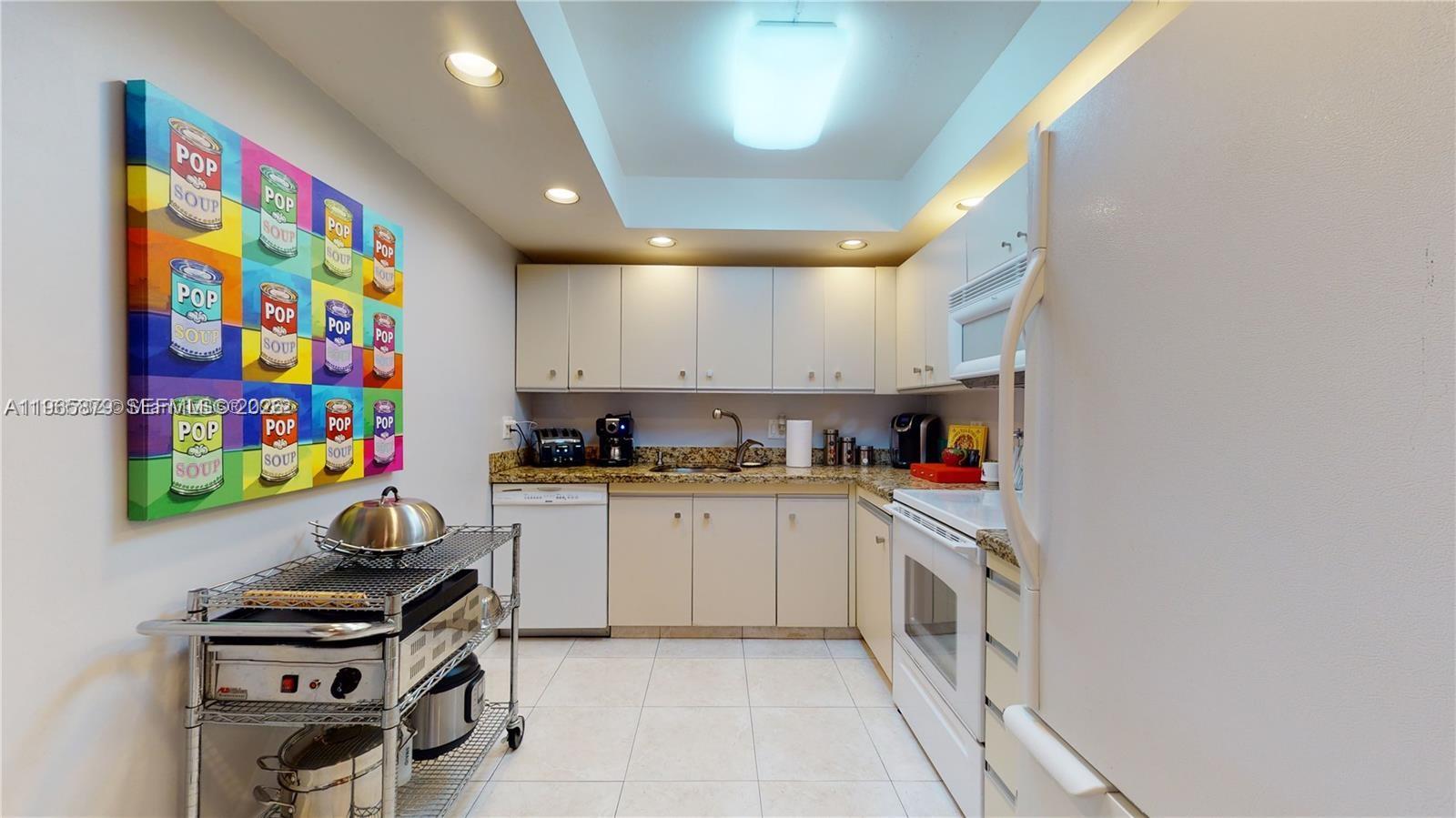 A galley kitchen features white cabinets, granite countertops, tile flooring, and a pop art print — 2500 PARKVIEW DR # 417, Hallandale Beach FL 33009, HALLANDALE BEACH, FL