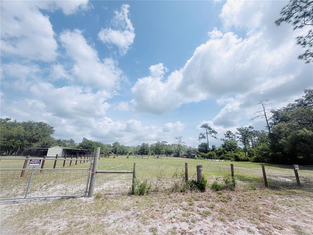 A fenced, grassy lot with a metal gate and scattered trees under a cloudy sky — TBD SW 153RD LANE, DUNNELLON, FL