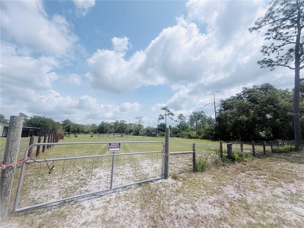 A metal gate opens onto a grassy, fenced-in lot with scattered trees under a cloudy sky — TBD SW 153RD LANE, DUNNELLON, FL