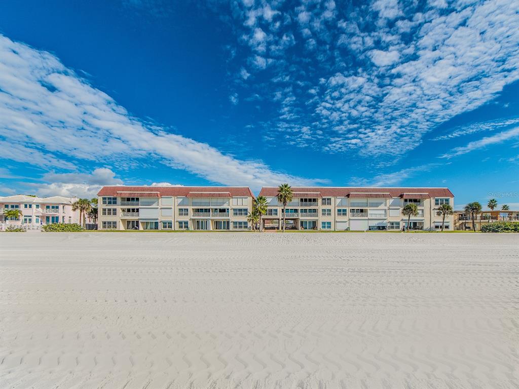 A multi-story beachfront apartment building sits behind a wide, sandy beach under a blue sky — 2900 GULF BOULEVARD UNIT 307, BELLEAIR BEACH, FL