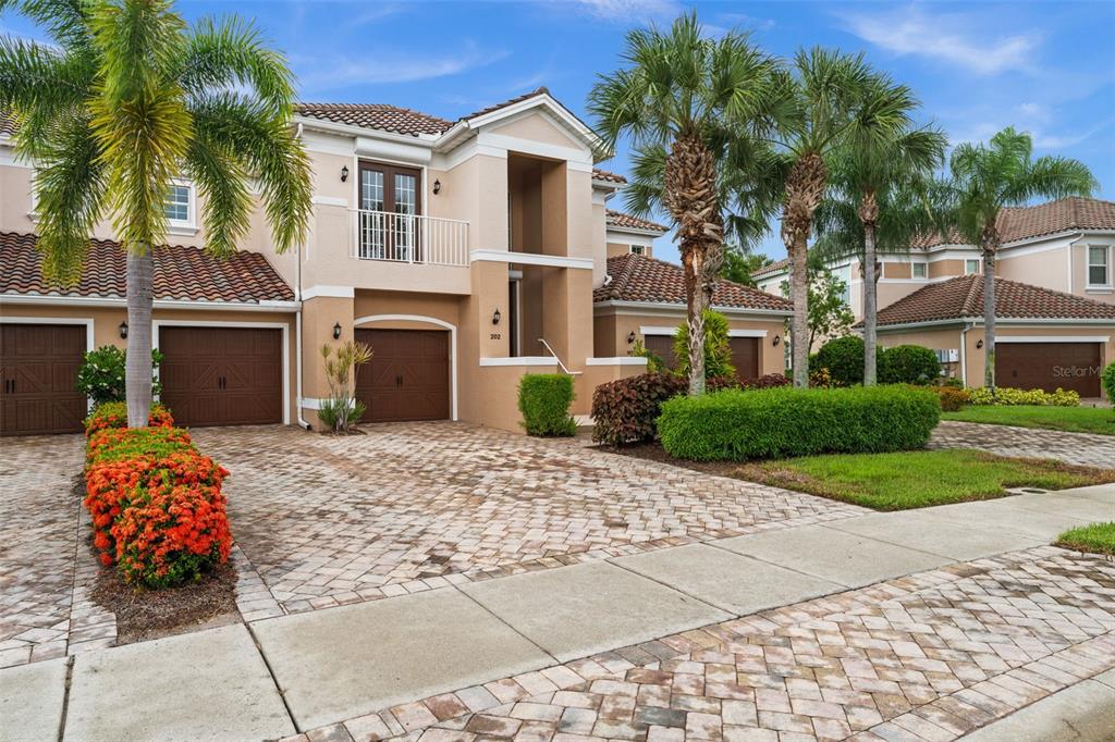 A two-story tan stucco townhouse with a paver driveway, brown garage doors, and palm trees — 10391 GLASTONBURY CIRCLE UNIT 202, FORT MYERS, FL
