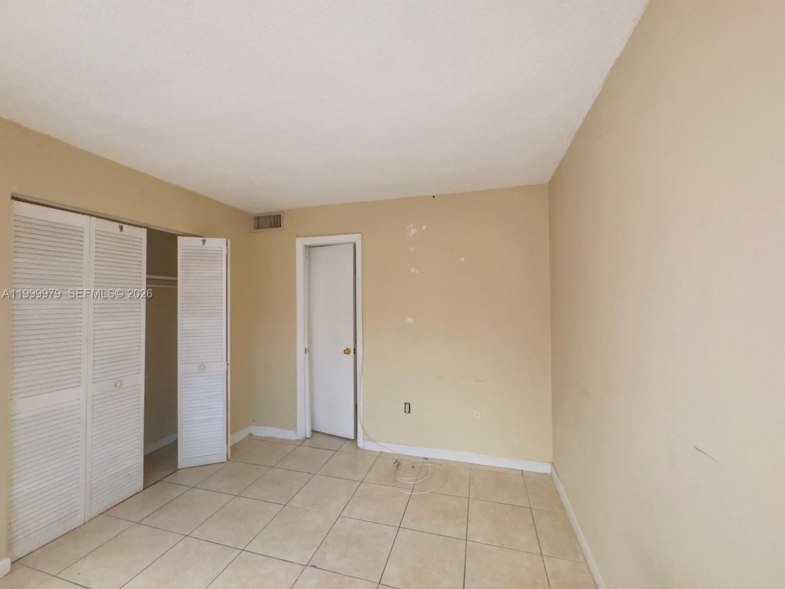 An empty bedroom features beige walls, white louvered closet doors, and light-colored tile flooring — 19841 SW 114th Ave # 310, Miami FL 33157, MIAMI, FL